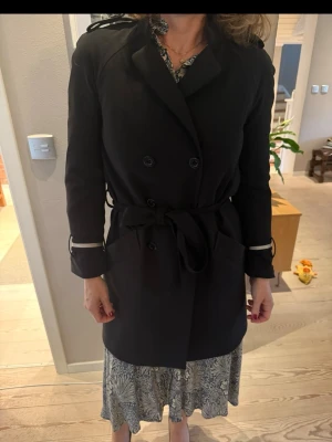 Svart trenchcoat från ALIX, S - Stilren svart trenchcoat från ALIX i storlek S. Jackan har dubbelknäppning, knytskärp i midjan och stora fickor framtill. Klassisk krage och dekorativa detaljer vid axlar och ärmslut. Perfekt för övergångssäsonger.