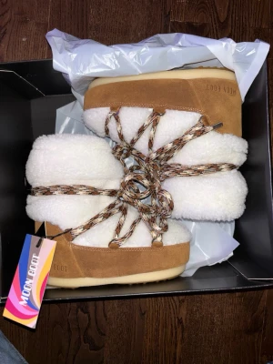 Moon boots low shearling storlek 33-34 - Moon Boot boots i vitt teddyfoder och brunt mocka med grova snören i beige och brunt mönster. Sulan är beige och gummiliknande med tydlig struktur för bra grepp. Perfekta för kalla dagar och riktigt mysiga att ha på sig.