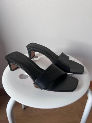 Svarta klackade sandaler från H&M - Stilrena svarta sandaler från H&M med fyrkantig tå och låg klack. Skorna har bred rem över foten och öppen häl, vilket ger en modern och minimalistisk look. Tillverkade i syntetmaterial med slät finish och passar perfekt till sommarens outfits. Använd en gång 