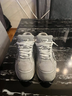 Dior B30 grå sneakers med CD-logga - Säljer ett par grå Dior B30 sneakers med stor CD-logga på sidan. Skorna har mesh och skinnpaneler, vita snören och en chunky vit sula. Snygg sportig vibe och riktigt clean design, perfekta för dig som gillar exklusiva märken och streetstyle.