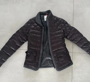 Mörkbrun quiltad dunjacka Moncler - Snygg mörkbrun dunjacka från Moncler med quiltad design och Moncler-logga på ärmen. Jackan har dragkedja, fickor med lock och grå detaljer vid krage och nederkant. 
