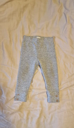Gråmelerade leggings med träknappar - Mysiga gråmelerade leggings med stickad känsla och två snygga träknappar på varje benslut. Perfekt för chill dagar och har en elastisk midja för skön passform. Stl 74.