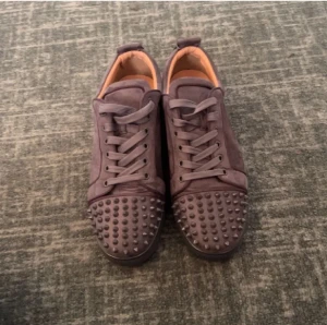 sneakers från Louboutin - Säljer ett par mörklila sneakers från Louboutin med ikoniska röda sulor och coola nitar på tån. Skorna har snörning, rund tå och är tillverkade i mocka med beige insida. Perfekt för dig som vill sticka ut med lyxig streetstyle.