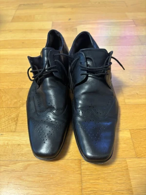 Bugatti kostym skor - Snygga svarta loafers från Ecco i äkta skinn med klassisk snörning och dekorativa perforeringar. Skorna har en platt sula och en elegant, spetsig tåform som ger en stilren look. Perfekta för dig som gillar dressad stil och vill ha något som sticker ut. Storlek 46 bra skick 