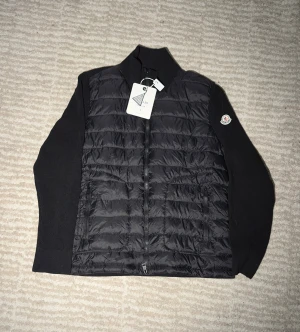 Svart Moncler pufferjacka med stickade ärmar - Svart pufferjacka från Moncler med quiltad front i glansigt material och stickade ärmar. Jackan har dragkedja framtill, ståkrage och Moncler-logga på vänster ärm. Perfekt för dig som vill ha en stilren och sportig look.