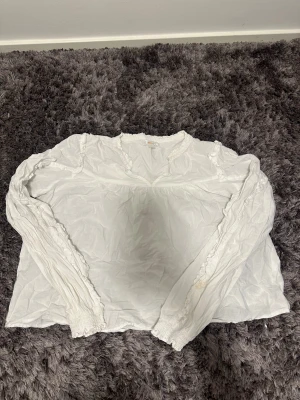 Vit blus med volang från Gina Tricot - Supersöt vit blus från Gina Tricot i storlek 170. Blusen har långa ärmar med smock vid ärmslut och fina volangdetaljer längs axlar och halsringning. Tillverkad i ett tunt, lätt tyg som känns luftigt och skönt.