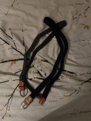 Två flätade bälten med skinn-detaljer - Säljer två snygga flätade bälten, ett i mörkgrått och ett i marinblått. Båda har bruna skinn-detaljer vid spännet och silverfärgat metallspänne. Perfekt för att ge outfiten en clean och stilren touch.