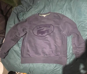 Mörkblå sweatshirt från Lacoste - Snygg mörkblå sweatshirt från Lacoste i storlek M. Tröjan har ett stort Lacoste-tryck med krokodil på bröstet och ribbade muddar vid ärmslut och nederkant. Perfekt för dig som gillar klassisk streetwear med en sportig touch.