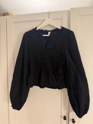 Svart blus från Nelly med smock - Svart blus från Nelly med smockdetaljer över hela livet och ärmarna. Blusen har långa puffärmar, knappar framtill och en lätt rynkad krage. Tillverkad i bomull för en luftig känsla.