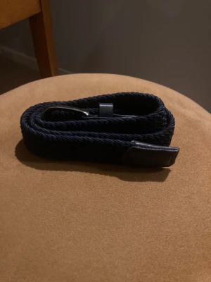 Mörkblå flätad bälte från one wear - Snyggt mörkblått flätat bälte med detaljer i skinn och metallspänne. Perfekt för att ge din outfit en stilren touch. Bältet har en klassisk form och passar till både jeans och chinos. Flätad design gör det enkelt att justera längden.