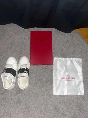 Valentino Garavani vita sneakers - Snygga vita sneakers från Valentino Garavani med bred marinblå rem över mitten och klassisk snörning. Skorna har rund tå, platt sula och är tillverkade i skinn. Perfekt för dig som gillar stilrena designer med en lyxig touch. Vid frågor kom dm! Inga skador på skorna! En påse o ena sidan till boxen ingår då den andra halvan har gick sönder! 