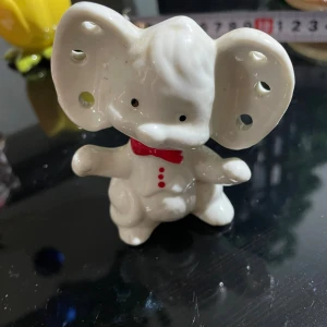 Söt elefantformad porslinsfigur - Charmig vit porslinsfigur i form av en elefant med stora öron och röda detaljer som fluga och knappar. Figuren har hål i öronen och botten, vilket gör den unik och lekfull. Perfekt som dekoration eller samlarobjekt.