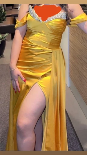 Gul långklänning med strassdetaljer - Säljer en snygg gul långklänning med offshoulder-ärmar och glittriga strassdetaljer vid bysten. Klänningen har en hög slits på sidan och är gjord i ett glansigt satinliknande material som ger en lyxig känsla. Perfekt för dig som vill sticka ut och känna dig extra glam.