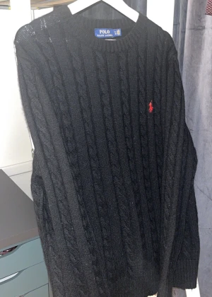 Svart kabel stickat ralph lauren tröja sweater - Svart stickad tröja från Polo Ralph Lauren i storlek XL. Tröjan har ett klassiskt kabelstickat mönster och en röd broderad logga på bröstet. Rund hals och långa ärmar med ribbade muddar. Perfekt för dig som gillar stilrena och tidlösa plagg.
