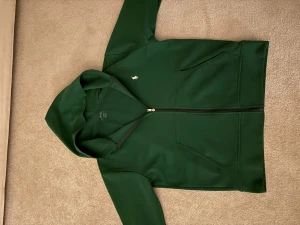 Grön hoodie från Polo Ralph Lauren - Snygg mörkgrön hoodie från Polo Ralph Lauren med dragkedja och huva. Tröjan har två fickor fram och den klassiska vita Polo-loggan broderad på bröstet. Pris kan diskuteras