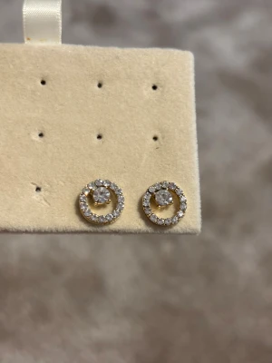 Guldiga örhängen med strass - Säljer ett par snygga runda örhängen i guldfärgad metall med glittrande strass-stenar runt hela cirkeln och en större sten i mitten. Perfekt för dig som gillar bling och vill ha något som sticker ut. Stift på baksidan för enkel användning.