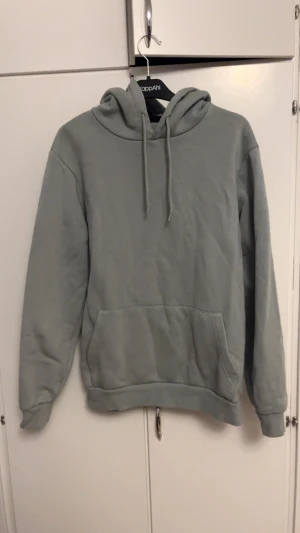 Grå hoodie från SMOG med huva - Snygg grå hoodie från SMOG i storlek M. Tröjan har en klassisk huva med snörning och en stor magficka framtill. Tillverkad i mjuk bomullsmix som är perfekt för chill dagar. Passformen är normal och ärmarna är långa för extra komfort.