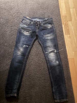 Dondup jeans -mörkblå distressed - Säljer ett par mörkblå dondup jeans med slitna detaljer  på benen. Jeansen har klassisk femficksdesign, knäppning med knappar och ljusa tvättade partier för en cool look.