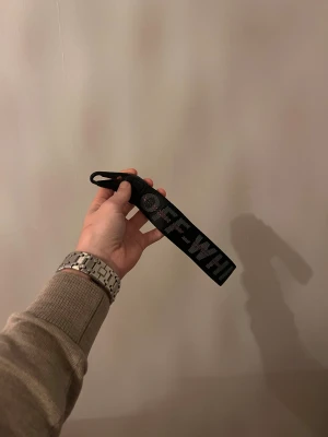 Svart Off-White nyckelring med zip tie - Säljer ett svart Off-White nyckelring med tydlig logotyp i grått och rött. Nyckelringen har en robust karbinhake och kommer med den ikoniska röda zip tie-taggen. Materialet är slitstark syntet och designen är bred och streetinspirerad, perfekt för att lyfta din outfit.