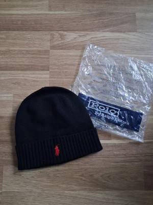 Svart stickad mössa från Polo Ralph Lauren - Svart ribbstickad mössa från Polo Ralph Lauren med klassisk röd broderad logga framtill. Mjuk och skön passform, perfekt för kyliga dagar. Materialet är stickad och designen är enkel med uppvikt kant.