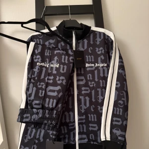 Palm angels sett  - Palm Angels sett med grått gotiskt mönster och vita ränder längs ärmarna. Tröjan har dragkedja framtill, ribbade muddar och klassisk krage. Materialet är polyester och tröjan har en loose passform. Perfekt för dig som gillar streetwear och vill sticka ut.