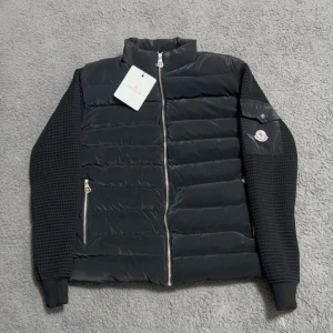 Svart Moncler pufferjacka  - Svart pufferjacka från Moncler med stickade ärmar och quiltad front. Jackan har dragkedja framtill, två fickor med dragkedja och en Moncler-logga på ärmen. Materialmix med polyester och stickad struktur på ärmarna ger en modern vibe. Storlek S men passar M!!