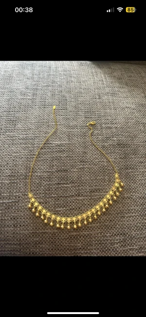 Guldigt halsband med hängande kulor - Unikt halsband i guldton med dekorativa små kulor som hänger längs hela kedjan. Kedjan har ett detaljerat, nästan blommigt mönster och ger ett riktigt statement till din look. Perfekt för dig som vill sticka ut med accessoarer som känns både trendiga och klassiska.