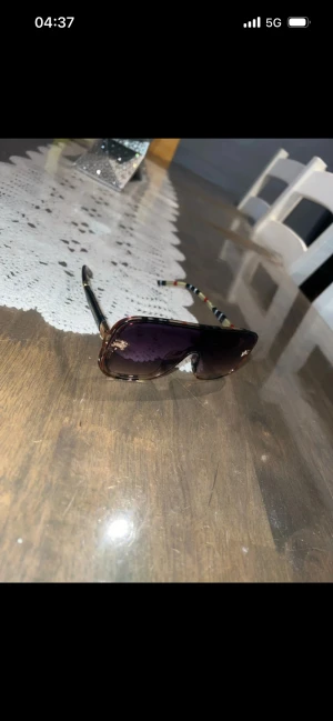 Burberry solglasögon med rutiga skalmar - Snygga Burberry solglasögon med stora, fyrkantiga bågar i brunmelerad plast och lila tonade glas. Skalmarna har klassiskt rutigt Burberry-mönster i rött, svart och beige samt gulddetaljer. Perfekt accessoar för att lyfta din stil.