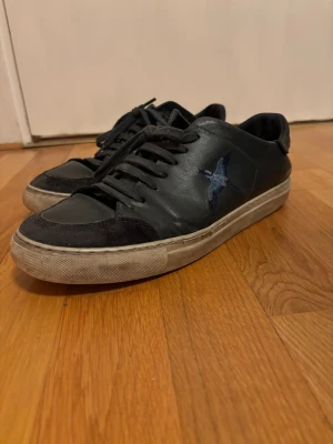 Svarta sneakers från Arigato med broderad fågel - Svarta sneakers från Arigato med snygg broderad fågel i blå toner på sidan. Skorna har snörning, rund tå och en vit platt sula. Ovandelen är i skinn med detaljer i mocka vid tån. Perfekt för dig som gillar stilrena och coola sneakers.