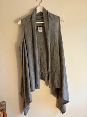 Grå omlottväst från Gina Tricot - Säljer en snygg grå omlottväst från Gina Tricot i modellen Wilda. Västen har en avslappnad passform med långa, draperade framstycken och är tillverkad i mjuk akryl. Perfekt att slänga över en t-shirt för en chill look.