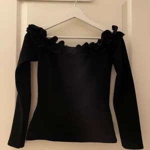 Svart offshoulder topp med volang - Säljer en svart offshoulder topp från ONLY TREND i storlek S. Toppen har långa ärmar och en bred volang längs halsringningen som ger en extra snygg touch. Perfekt för dig som gillar trendiga detaljer och stilren design.