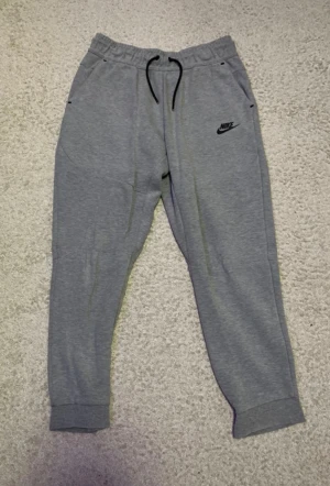 Grå Nike tech byxa storlek L Juniorstorlek - Grå mjukisbyxor från Nike med svart logga på vänster lår. Byxorna har elastisk midja med snörning, ribbade muddar vid benslut och praktiska fickor, varav en med dragkedja bak. Perfekta för chill eller träning.