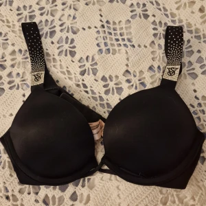 Svart push-up bh Victoria's Secret - Svart push-up bh från Victoria's Secret med vadderade kupor och axelband dekorerade med små silvriga prickar. Snygg och stilren design, perfekt för dig som vill ha extra lyft. Mjukt material och bekväm passform. Kostar lite extra pga att det står VS på axelbanden. Säljer för att jag råkade köpa fel storlek och är alldeles för liten. Använd 1 gång!😊