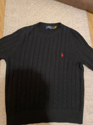 Svart stickad tröja Polo Ralph Lauren - Svart kabelstickad tröja från Polo Ralph Lauren med klassisk rund hals och röd broderad logga på bröstet. Långärmad modell i mjukt stickat material som ger en clean och tidlös vibe. Perfekt för dig som gillar stilrena plagg med lite preppy känsla.