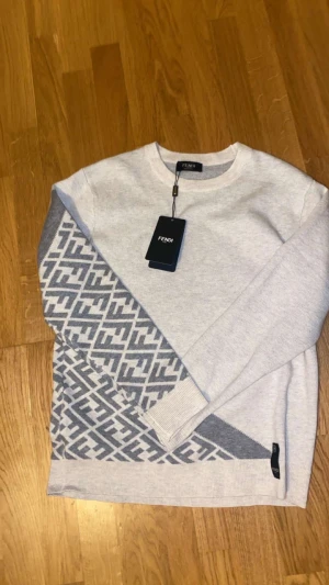 Grå stickad Fendi tröja med mönster - Snygg ljusgrå stickad tröja från Fendi med rund hals och lång ärm. Tröjan har ett stort, diagonalvänt Fendi-logomönster i mörkgrått på ena sidan och ribbade muddar. Perfekt för dig som gillar exklusiva märken och stilrena detaljer.