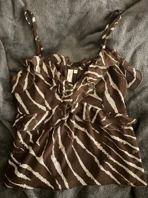 Brun zebramönstrad volangtop från Nelly - Säljer en brun och vit zebramönstrad topp från Nelly i storlek 34. Toppen har tunna axelband, v-ringning och flera volangdetaljer framtill och upptill. Materialet är lätt och luftigt, perfekt för varma dagar. Supersnygg att styla med jeans eller kjol!