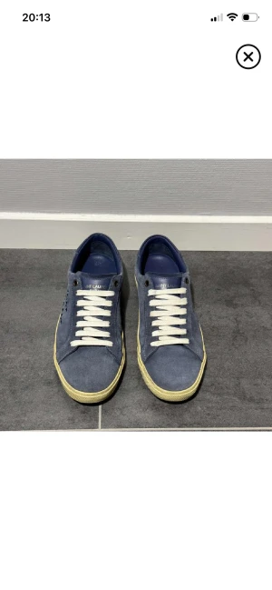 Blå Saint Laurent sneakers mocka - Snygga blå sneakers från Saint Laurent med handskriven logga på sidan. Skorna har vit snörning, beige platt sula och är tillverkade i mjuk mocka med blå detaljer vid hälen. Perfekt för dig som gillar stilrena och exklusiva sneakers.