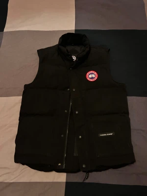 Svart dunväst från Canada Goose - Svart dunväst från Canada Goose med hög krage, två stora fickor framtill och klassisk logopatch på bröstet. Västen har både dragkedja och tryckknappar, samt märkning på fickan. Perfekt för lager-på-lager under kyliga dagar.