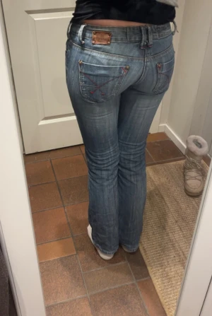 Vintage Lågmidjade bootcut jeans  - Säljer ett par blå jeans med låg midja och bootcut-snitt. Jeansen har klassisk femficksdesign, röda broderade detaljer på bakfickorna och en läderpatch med logga bak. Materialet är slitstarkt denim i bomull med snygg tvättad look.