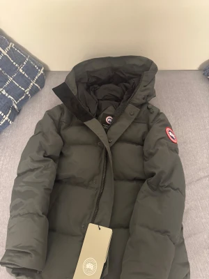 Macmilian Canada goose jacka  - Säljer en Grå pufferjacka från Canada Goose med ikonisk patch på armen och huva. Jackan är dunfylld och har dragkedja framtill. Perfekt för kalla dagar och riktigt snygg streetstil.