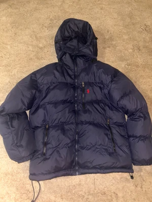 Mörkblå dunjacka från Polo Ralph Lauren - Snygg mörkblå dunjacka från Polo Ralph Lauren med huva och dragkedjor framtill. Jackan har det klassiska röda Polo-logot broderat på bröstet, två sidofickor med dragkedja och justerbar nederkant. Perfekt för kalla dagar och riktigt chill stil.L