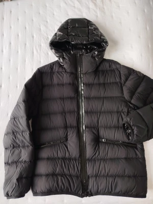 Svart Moncler pufferjacka med huva - Säljer en svart pufferjacka från Moncler med glansig finish och huva. Jackan har två sidofickor med dragkedja, quiltad design och Moncler-logga på ärmen. Perfekt för kalla dagar och har en snygg, modern look.