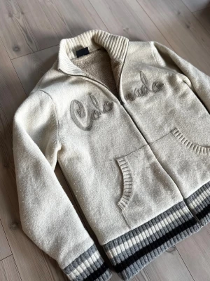 Unik teddycardigan från Scotch & Soda - Mysig vit teddycardigan från Scotch & Soda med broderad 'Colorado'-text på bröstet. Jackan har ribbade muddar och nederkant i grått och vitt, dragkedja framtill och två fickor. Insidan är fodrad med mjuk teddy för extra värme och komfort. Storlek s/m