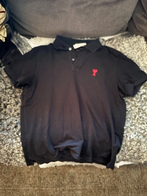 Svart piké från Ami med rött hjärta - Svart pikétröja från Ami med klassisk krage och knappar framtill. På bröstet finns ett snyggt rött broderat hjärta med bokstaven A. Materialet är mjuk bomull och modellen har korta ärmar. Perfekt för dig som gillar stilrena detaljer. Storlek M men passar även S. 100% äkta då den är köpt från nk 2023. Skriv vid minsta lilla fundering🙌🏼 Ny pris: 1700kr. Mitt pris: 699kr.