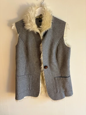 Grå pälsjacka från Vila, storlek S - Säljer en grå ärmlös pälsjacka från Vila med fluffig vit fuskpäls på insidan och kragen. Jackan har en snygg brun knapp framtill(saknar en knapp enligt bild 2) och två stora fickor. Perfekt för dig som vill ha en cool lager-på-lager look under höst och vinter. Saknar bältet.