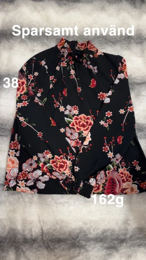 Svart blommig blus med hög krage - Supersnygg svart blus med färgglada blommor i rött, rosa och orange. Hög smockad krage och långa ärmar med smockade muddar. Luftigt tyg som känns lätt och flowy. Perfekt för dig som gillar statement-mönster och trendiga detaljer.🌸Sparsamt använd. Storlek 38