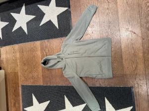 Grön hoodie från Sail Racing - Snygg nature green hoodie från Sail Racing med dragkedja och huva. Tröjan har två sidofickor med dragkedja och en stilren design. Perfekt för dig som gillar sportig och avslappnad stil. Materialet är mjukt och skönt, med fleece på insidan för extra komfort.