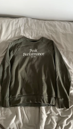 Grön Peak Performance sweatshirt - Snygg grön sweatshirt från Peak Performance med vit logotyp på bröstet. Tröjan har rund hals, långa ärmar och ribbade muddar vid ärmslut och nederkant. Perfekt för chill dagar och enkel att matcha med jeans eller joggers.