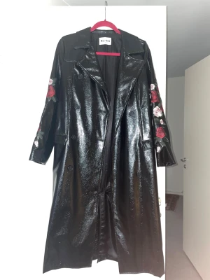 Svart trenchcoat med rosbroderi NA-KD - Säljer en svart glansig trenchcoat från NA-KD i storlek 34. Jackan har broderade rosor i rött och rosa på ärmarna, knappar framtill och bälte i midjan. Lång modell med klassisk krage och snyggt fall. Perfekt statement-plagg för dig som gillar att sticka ut.