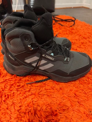 Adidas Terrex svarta kängor strl 38 - Grymma Adidas Terrex kängor i svart och grått med robust sula och snörning. Perfekta för utomhusaktiviteter, med slitstarkt syntetmaterial och klassiska tre ränder på sidan. Hög modell som ger bra stöd och skydd för foten.